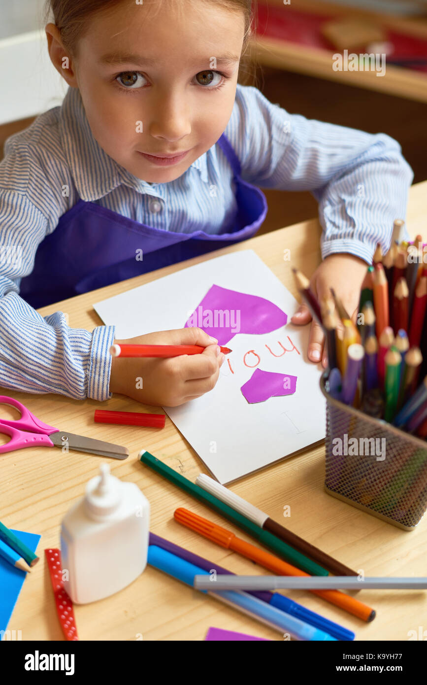 Portrait of adorable little girl looking up at camera while making gift card for mom during art class in pre-school Stock Photo