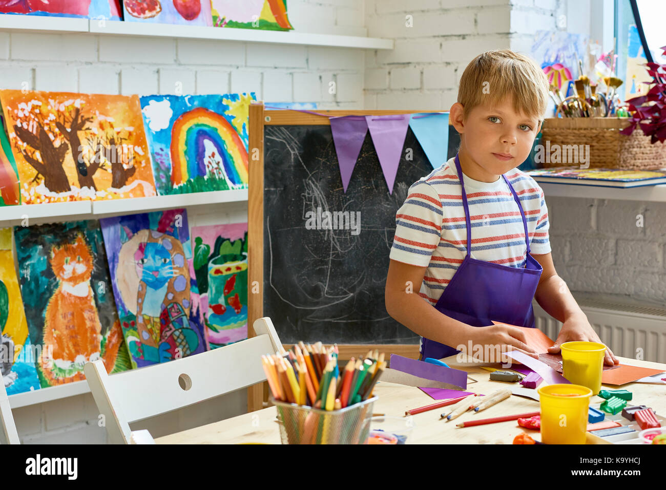 Portrait of blonde little boy looking at camera while making handmade gift card during art and craft lesson of pre-school, copy space Stock Photo