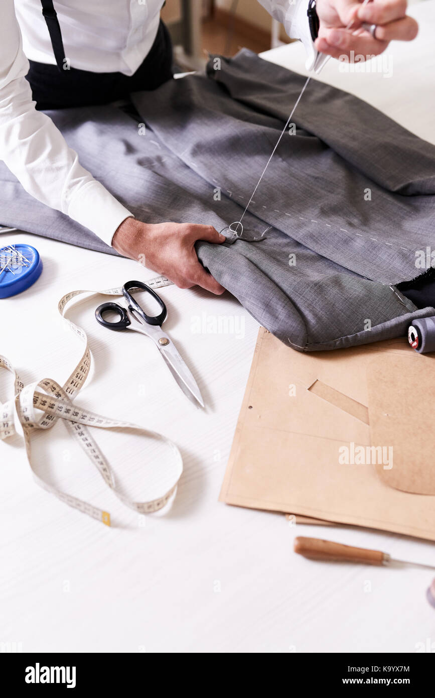 Highly professional young tailor working on custom-made male jacket with help of thread and needle while standing at table, close-up shot Stock Photo