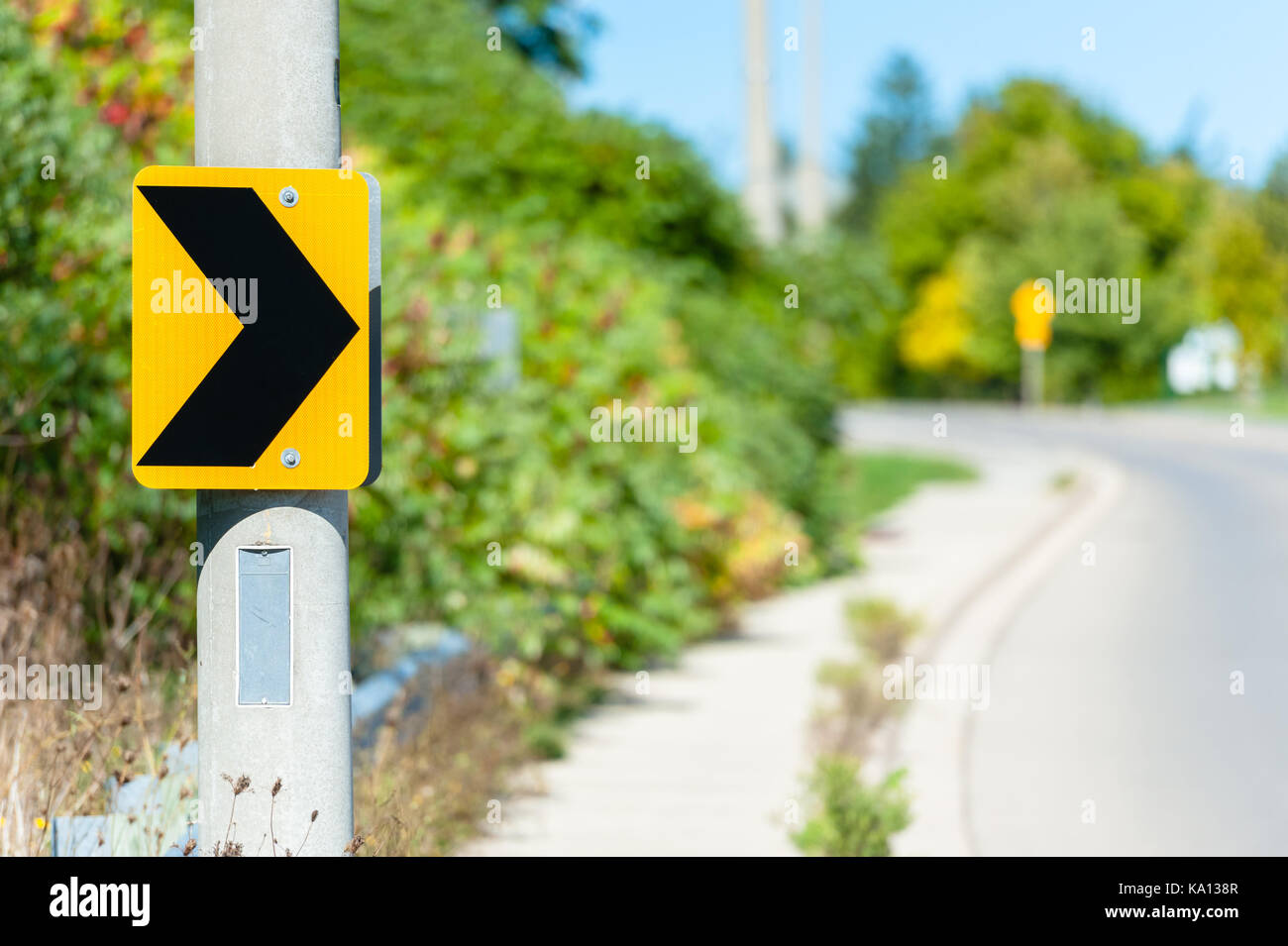 Black on yellow chevron road sign attached to post, indicating right turn against blurry curving road in background. Stock Photo