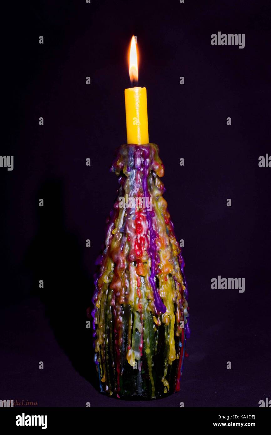 Yellow candle lit on a bottle used as a candlestick, the melted wax from previously used candles makes up a history of use Stock Photo