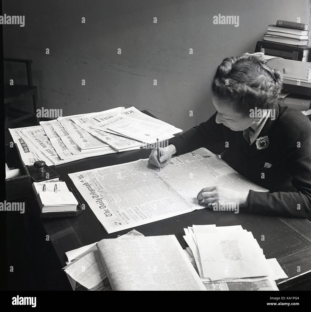 1950s, historical, well-dressed female working at a desk looking through the UK daily newspaper 'The Daily Telegraph' for articles of interest. On her desk she has other daily newspapers and publications. Stock Photo