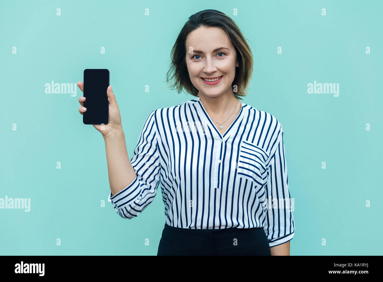 Success business woman showing new smart phone and looking at camera with toothy smile. Outdoor shot, blue background Stock Photo