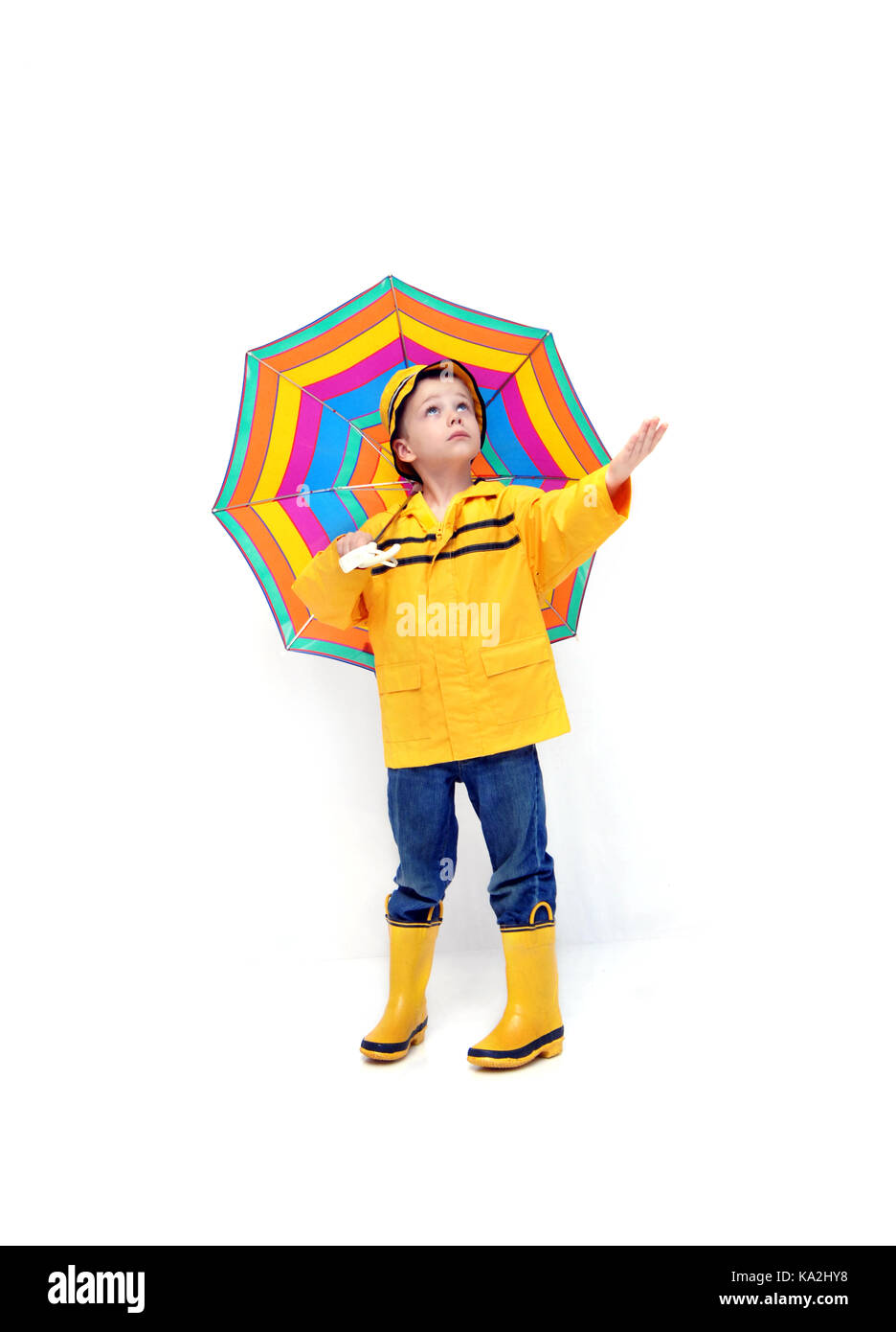 Young boy in yellow raincoat and rubber boots holds his hand out to check for rain.  He is holding a colorful striped umbrella. Stock Photo