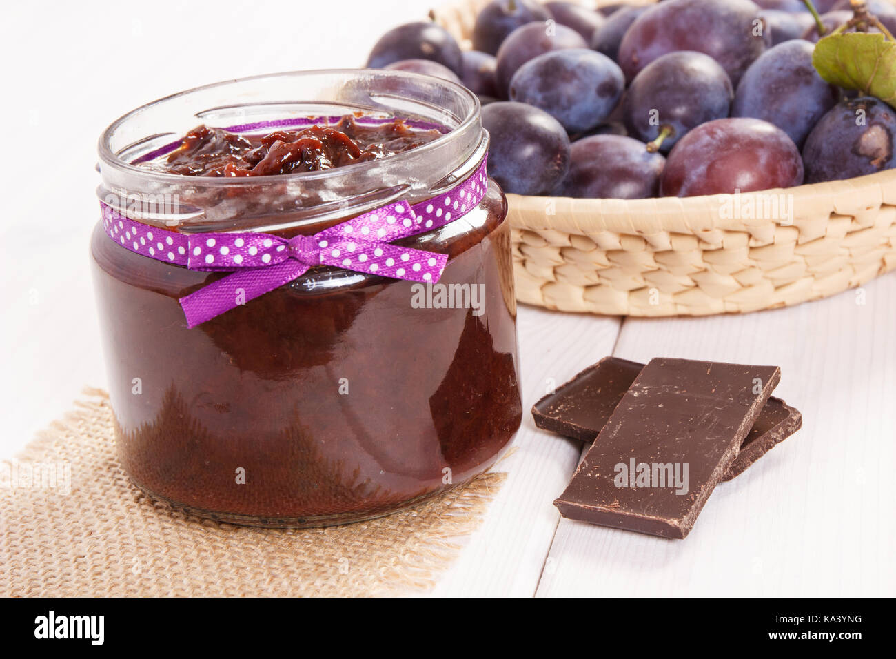 Fresh plum jam or marmalade in glass jar, ripe fruits in wicker basket and chocolate, concept of healthy sweet dessert Stock Photo