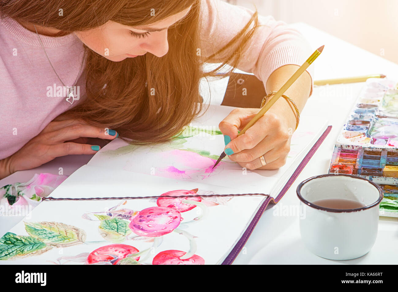 Dark-haired young woman draws a wooden brush and watercolor in a drawing pad with pink peonies with green leaves, on the table lie a leather case with Stock Photo