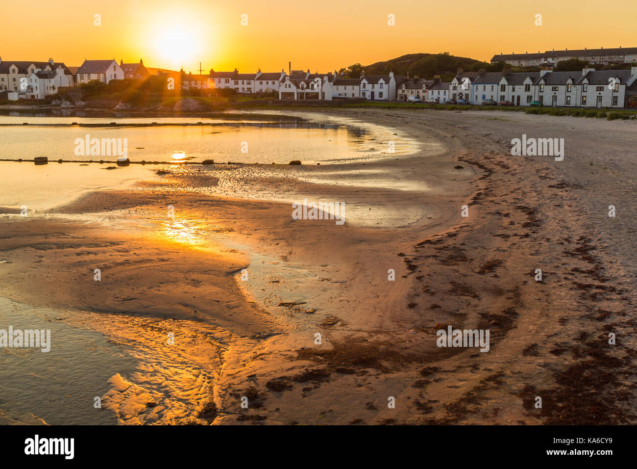 Town of Port Ellen, shot in direct light at evening time with the sandy  beach in front and houses in the background, sun making nice color in the  sand Stock Photo -