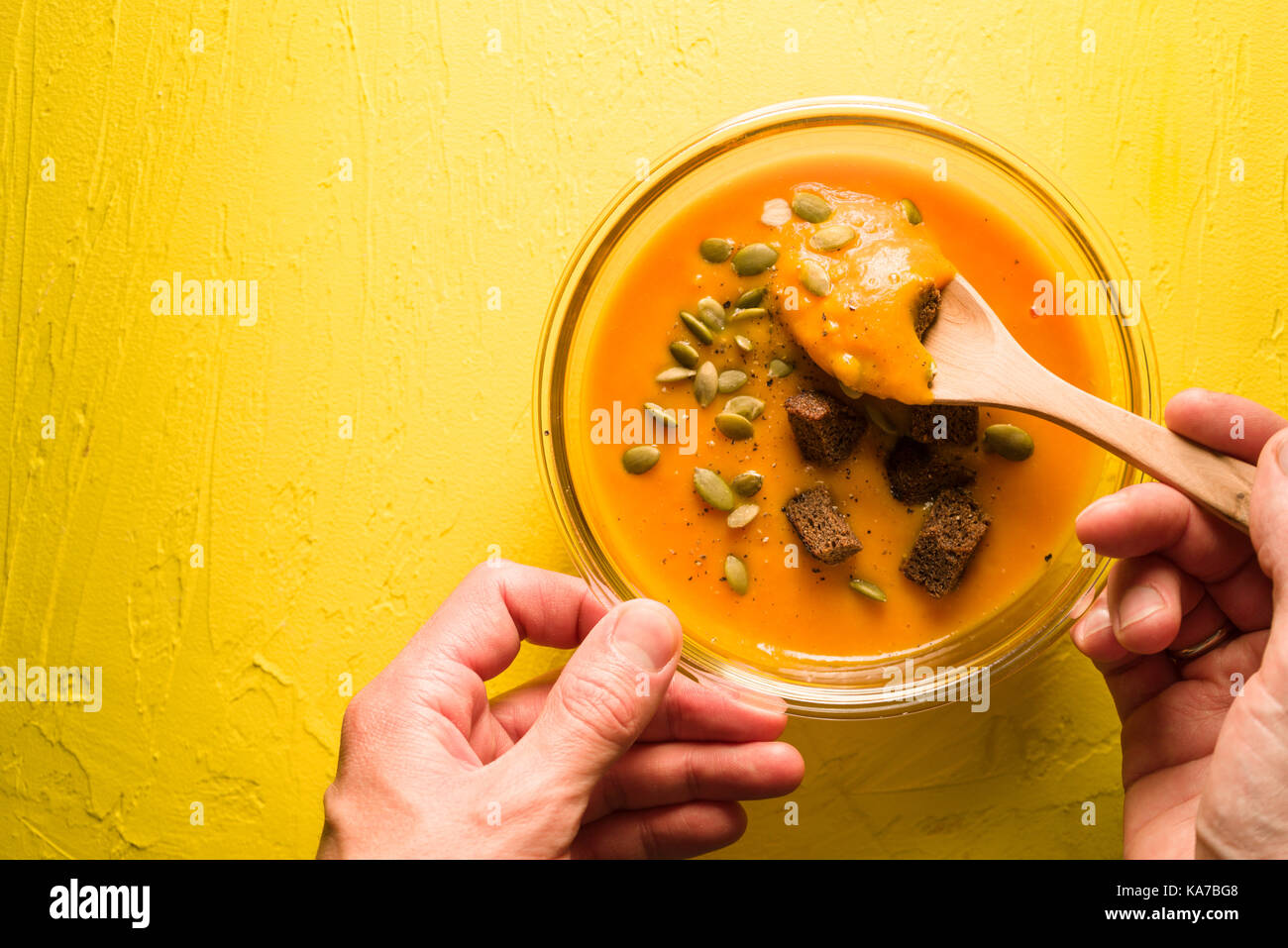 Pumpkin soup with seeds and croutons on a yellow table and spoon in hand horizontal Stock Photo
