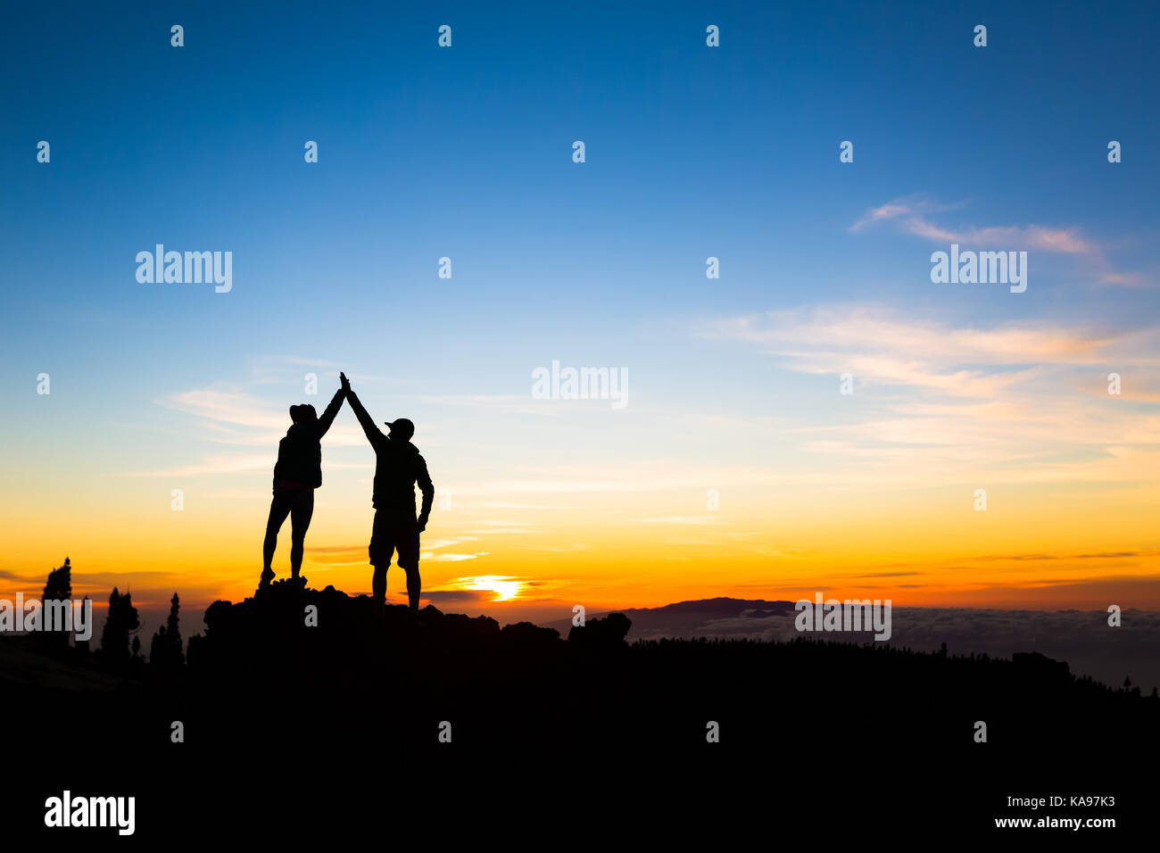 Couple hikers success in sunset mountains, accomplished with arms up outstretched. Man and woman on rocky mountain looking at beautiful sunset inspira Stock Photo