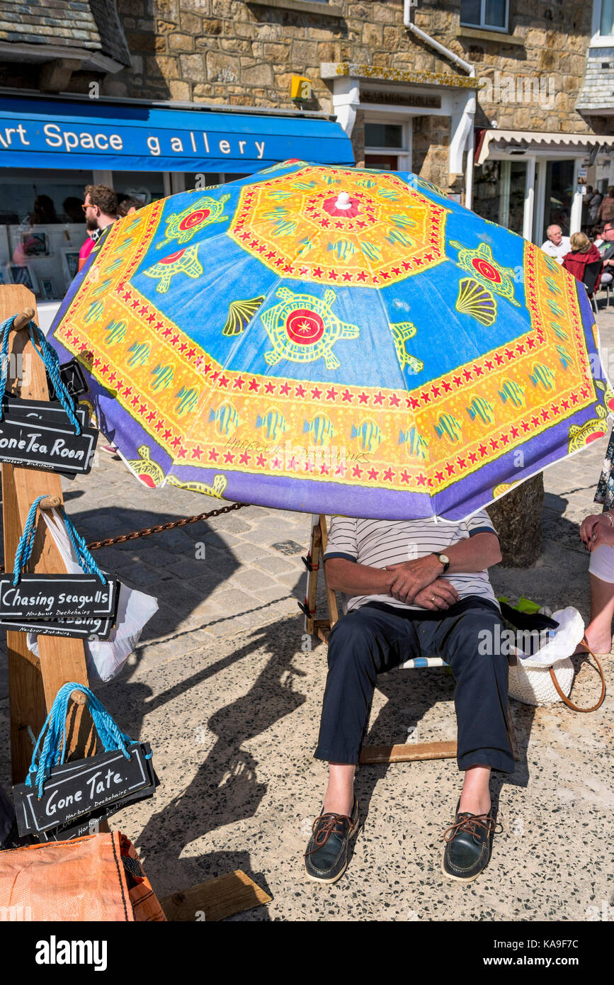 St Ives - a holidaymaker relaxing in the shade of large colourful umbrella near St Ives Harbour n St Ives in Cornwall. Stock Photo