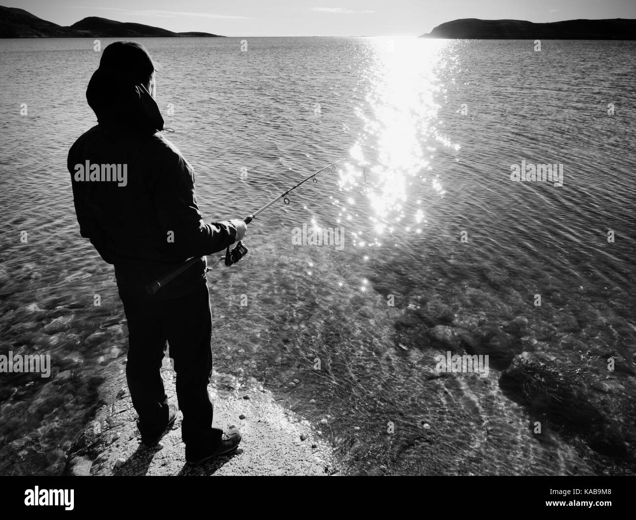 The silhouette of a fisherman who throws spinning at dawn . The active man is fishing on sea from the rocky coast. Stock Photo