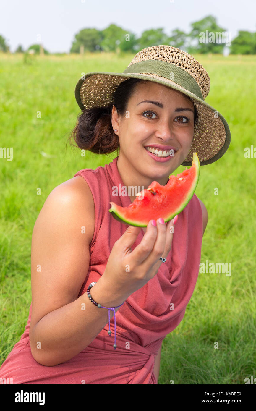 Young woman wearing straw hat eating fresh red melon in nature Stock