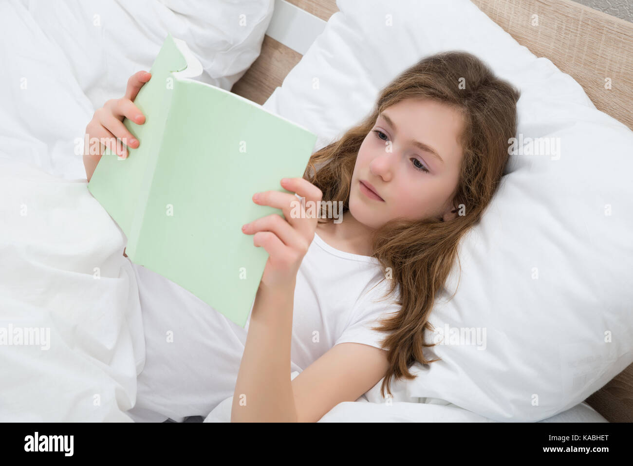 Cute Girl With Pillow Reading Book And Lying In Bed Stock Photo