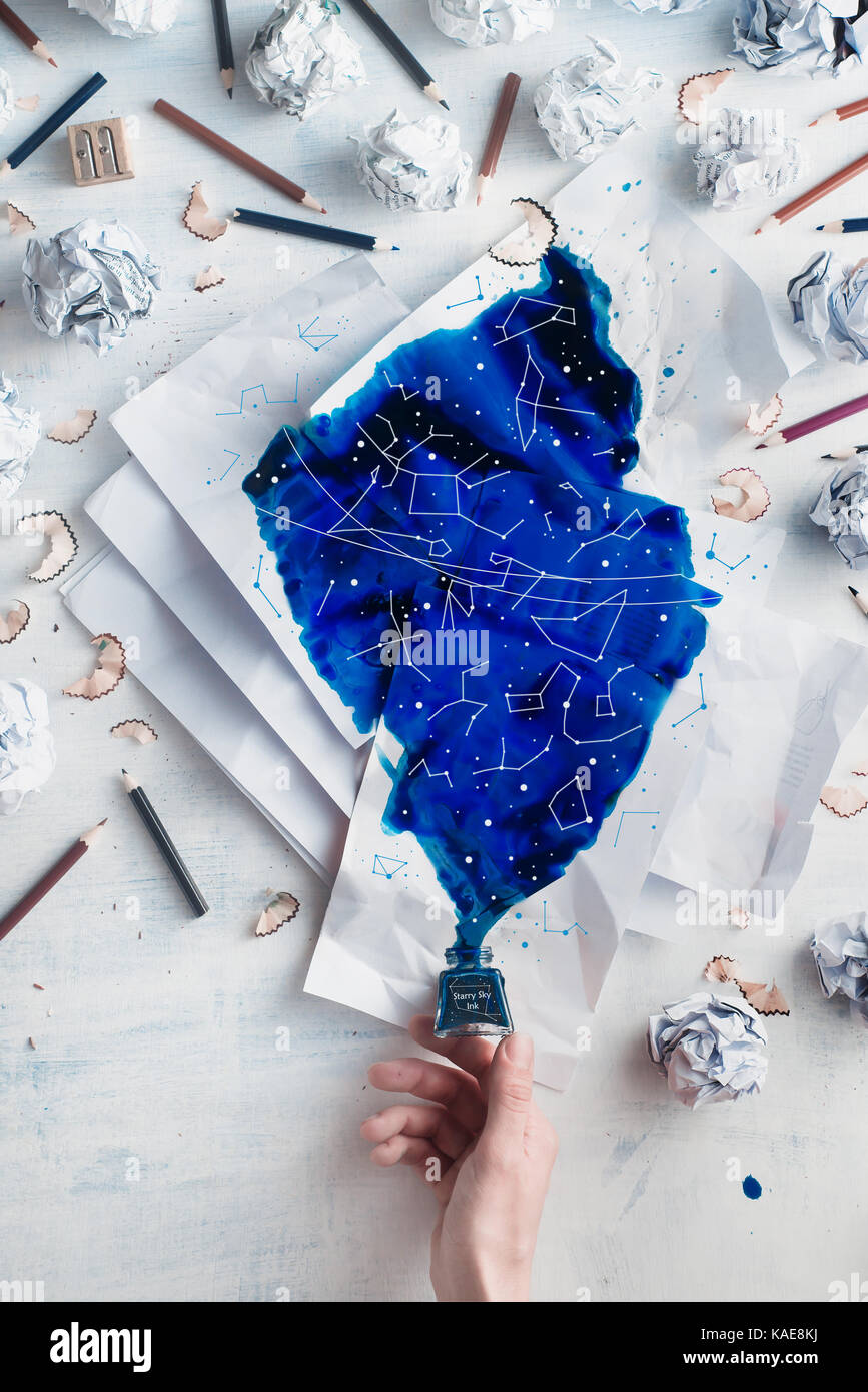 Crumpled paper balls with pencils and papers on a white wooden background with spilled ink forming starry sky with constellations. Writers hand holdin Stock Photo