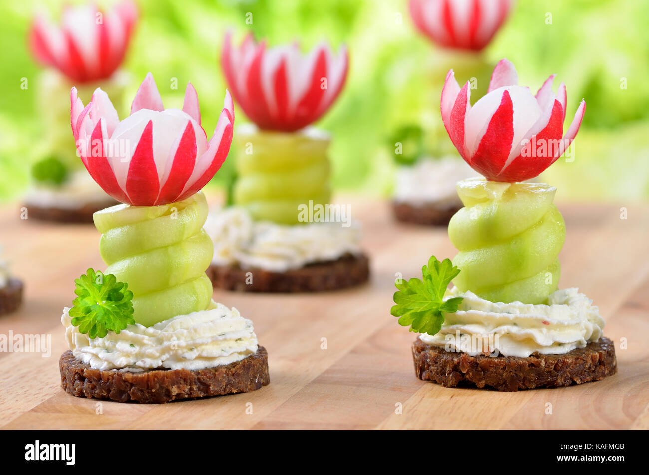 Decorated cheese appetizers: Spiced cream cheese with spiral cucumber and radishes blossoms on dark wholegrain bread Stock Photo
