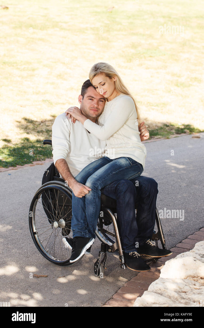Young girl sitting on the lap of her disabled boyfriend in a wheelchair in the park Stock Photo