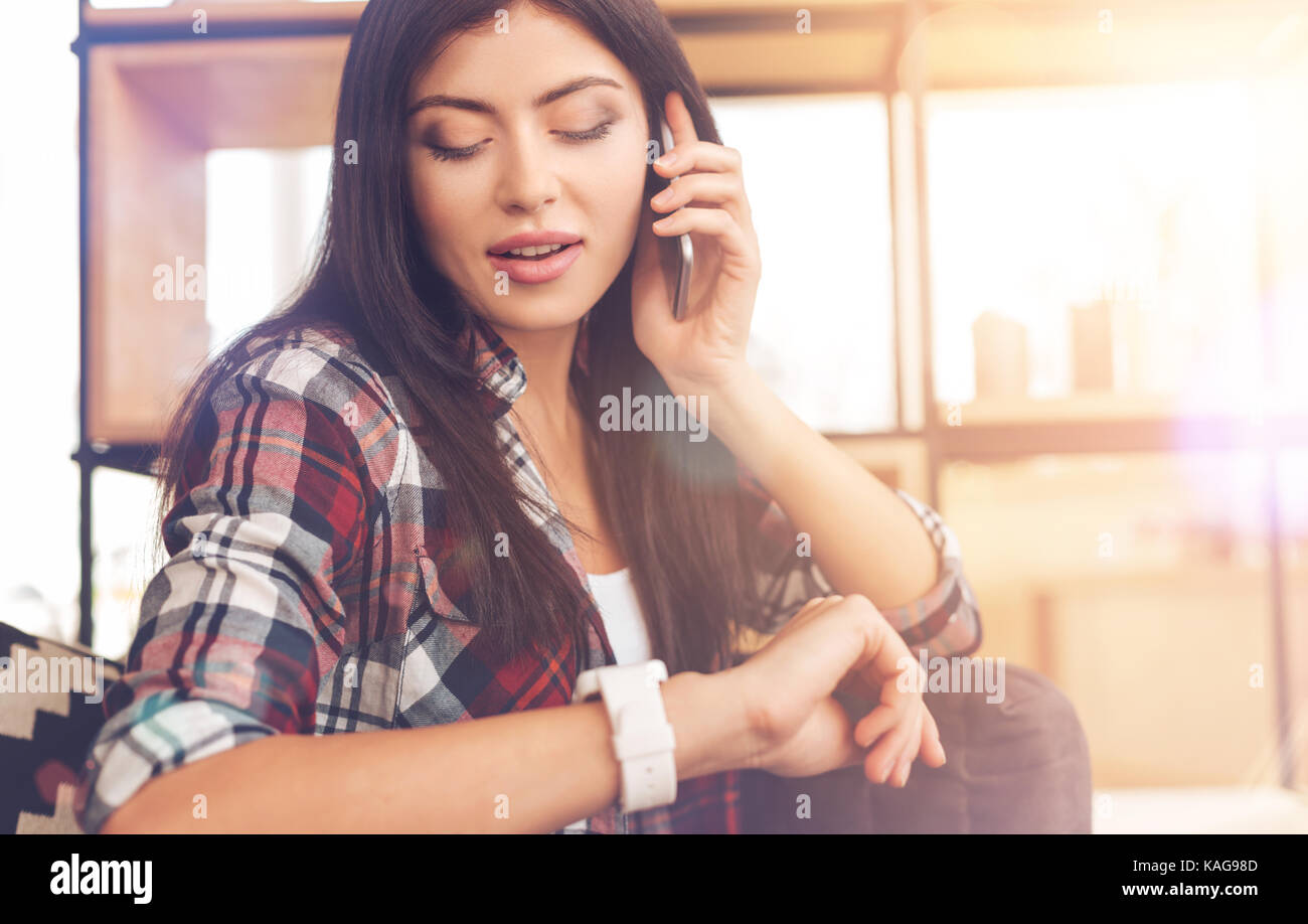 Time is money. Beautiful brunette wearing casual attire looking at a wrist watch for time checking while sitting on a sofa and having a serious busine Stock Photo