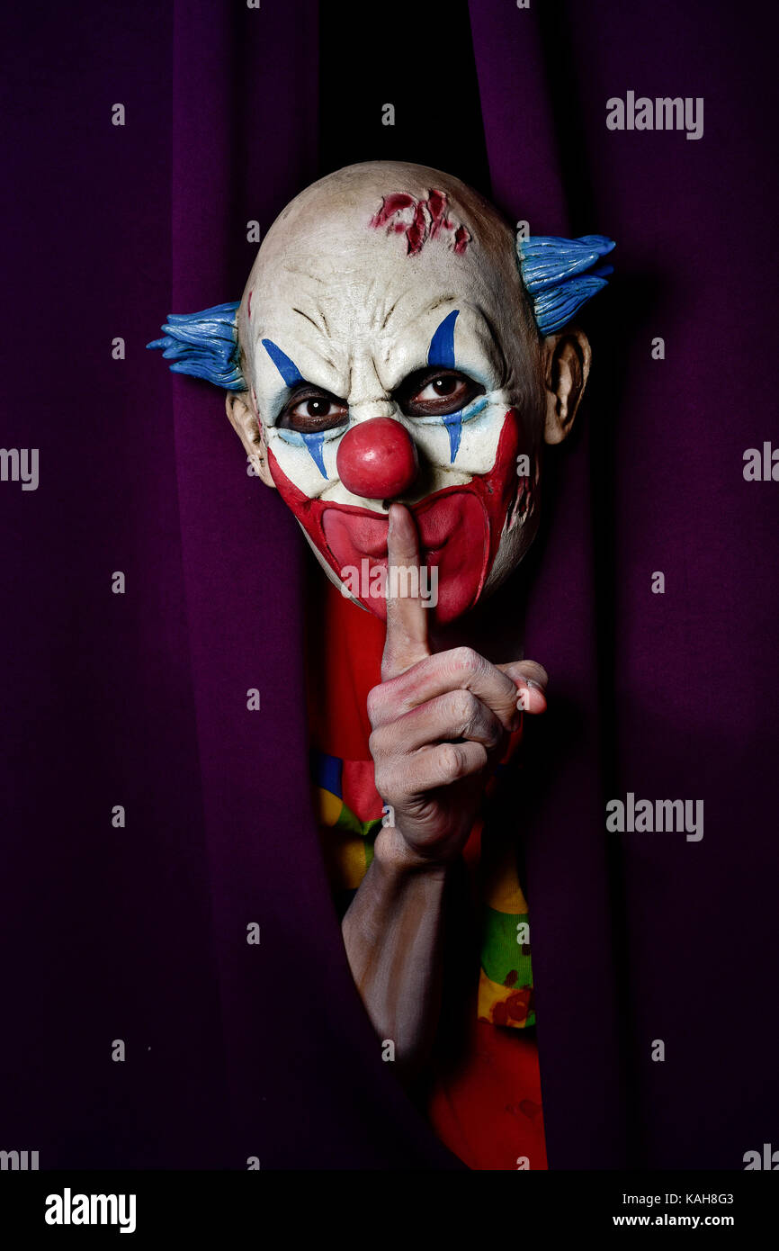 a scary evil clown peering out from a purple stage curtain, with his