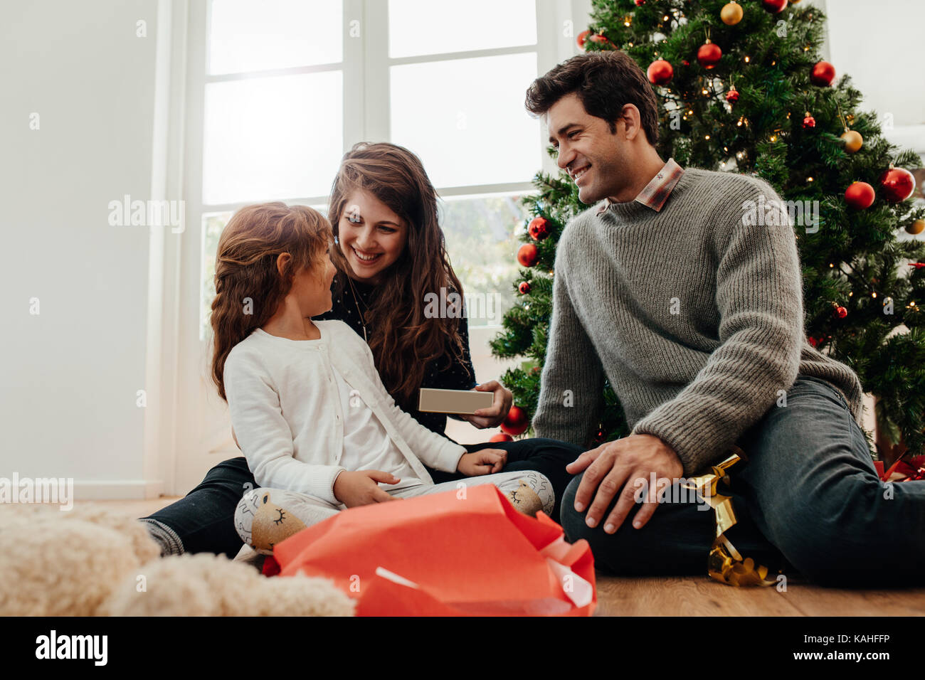 Family having happy time celebrating Christmas at home. Young couple with their daughter opening gifts sitting near Christmas tree. Stock Photo