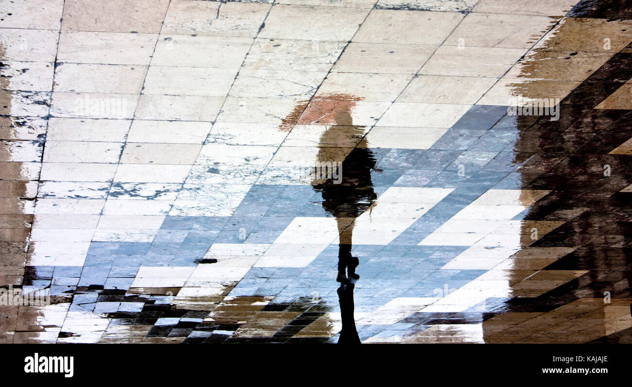 Blurry reflection shadow silhouette of a single woman under red umbrella walking in the old city pedestrian zone just after the rain Stock Photo