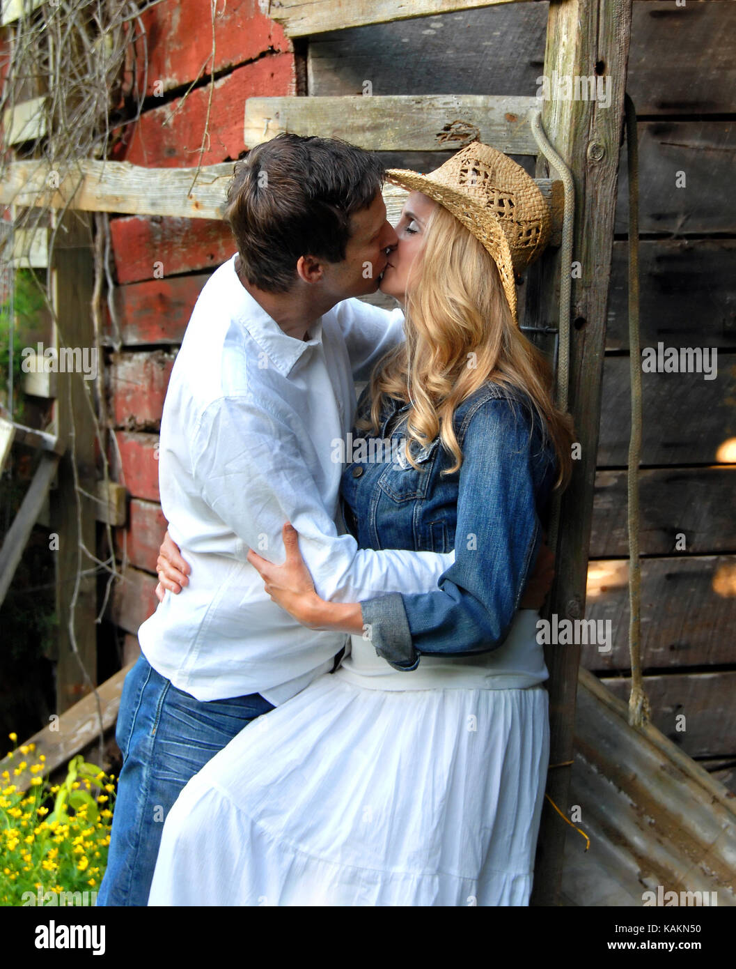 Couple share a romantic kiss leaning against a rustic, red wooden barn.  She is wearing a denim jacket and white dress.  He is wearing jeans and a whi Stock Photo
