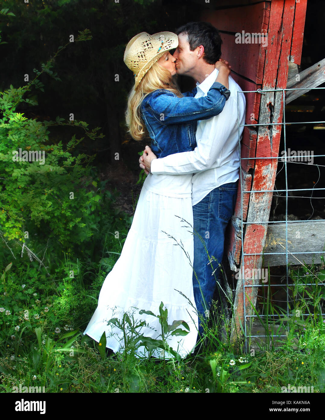 Young couple embrace behind the barn.  She is wearing a white dress with cowboy hat.  He is wearing jeans and a white shirtl Stock Photo