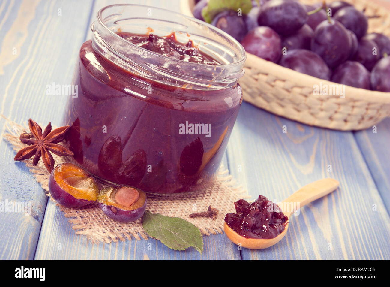 Vintage photo, Fresh homemade plum marmalade in glass jar, spices and ripe fruits in wicker basket in background, concept of healthy sweet dessert Stock Photo
