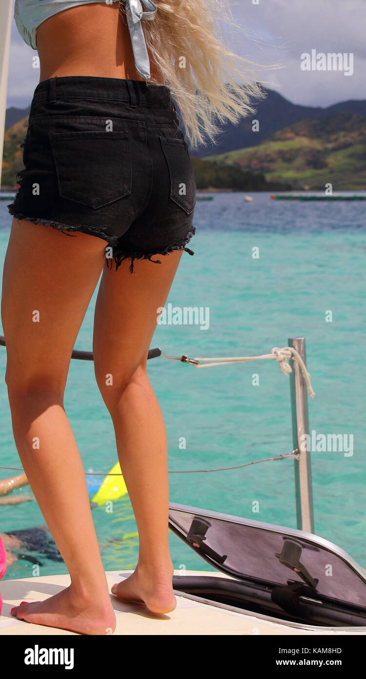 View from behind of sun tanned human legs on the deck of a yacht in portrait format with a tropical island background with copy space Stock Photo