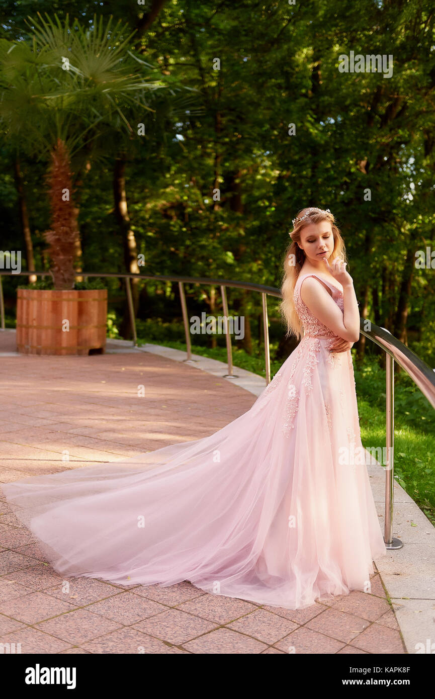 Portrait of a beautiful bride blond girl in pink lace dress ,hair decoration, handmade. Tenderness. Standing in the midst of a dark forest green in the rays of the setting sun.Shows a long dress with a train Stock Photo