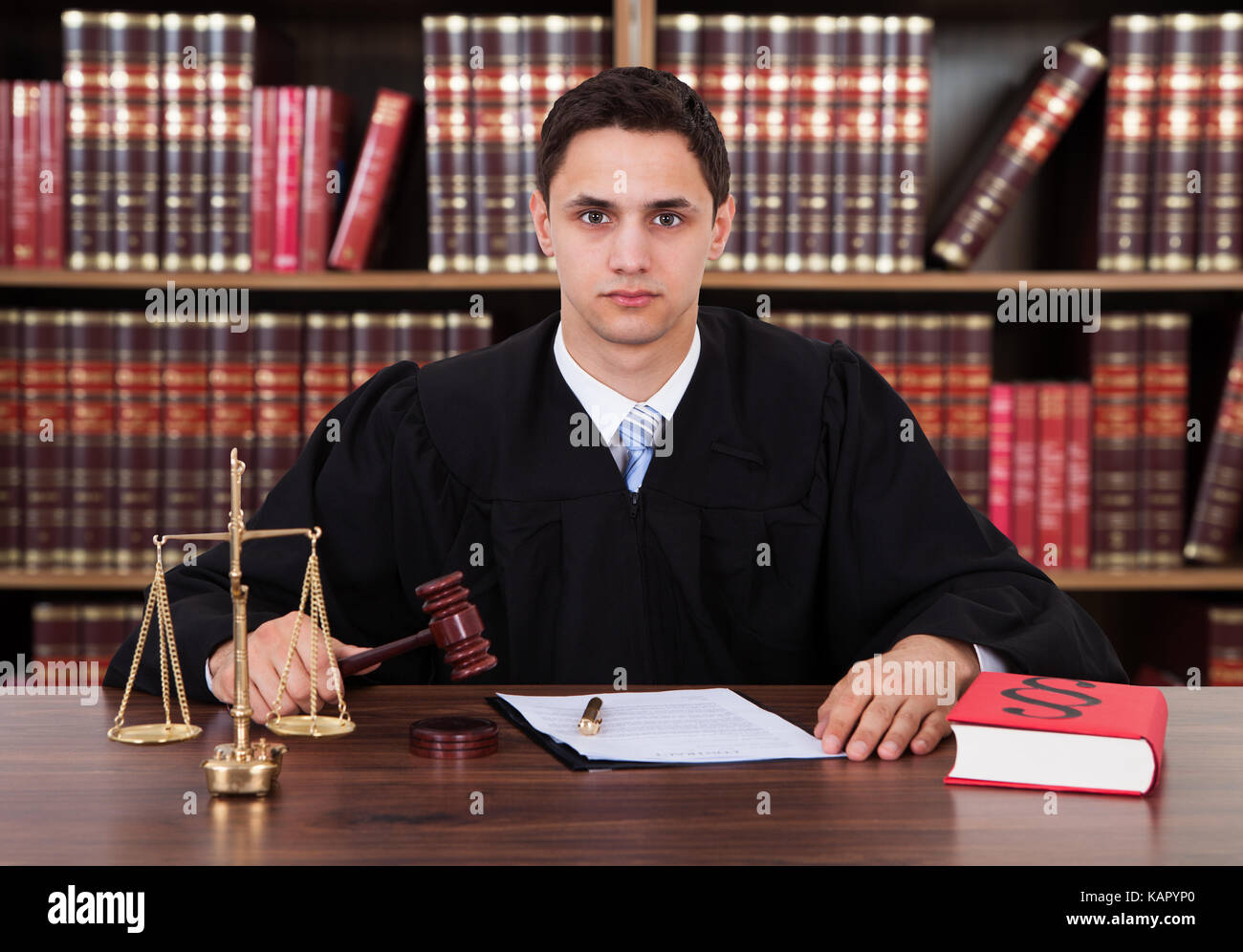 Portrait of confident young male judge striking the gavel at table against bookshelf Stock Photo