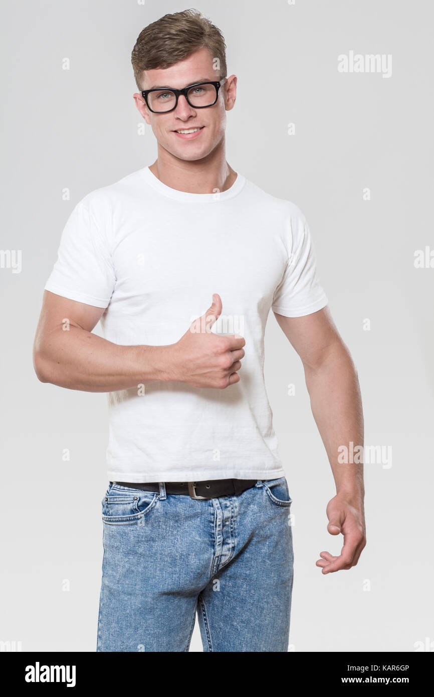 Smiling handsome young man with thumbs up. Picture taken in a studio with grey background. The model is wearing glasses and a white t-shirt. Stock Photo