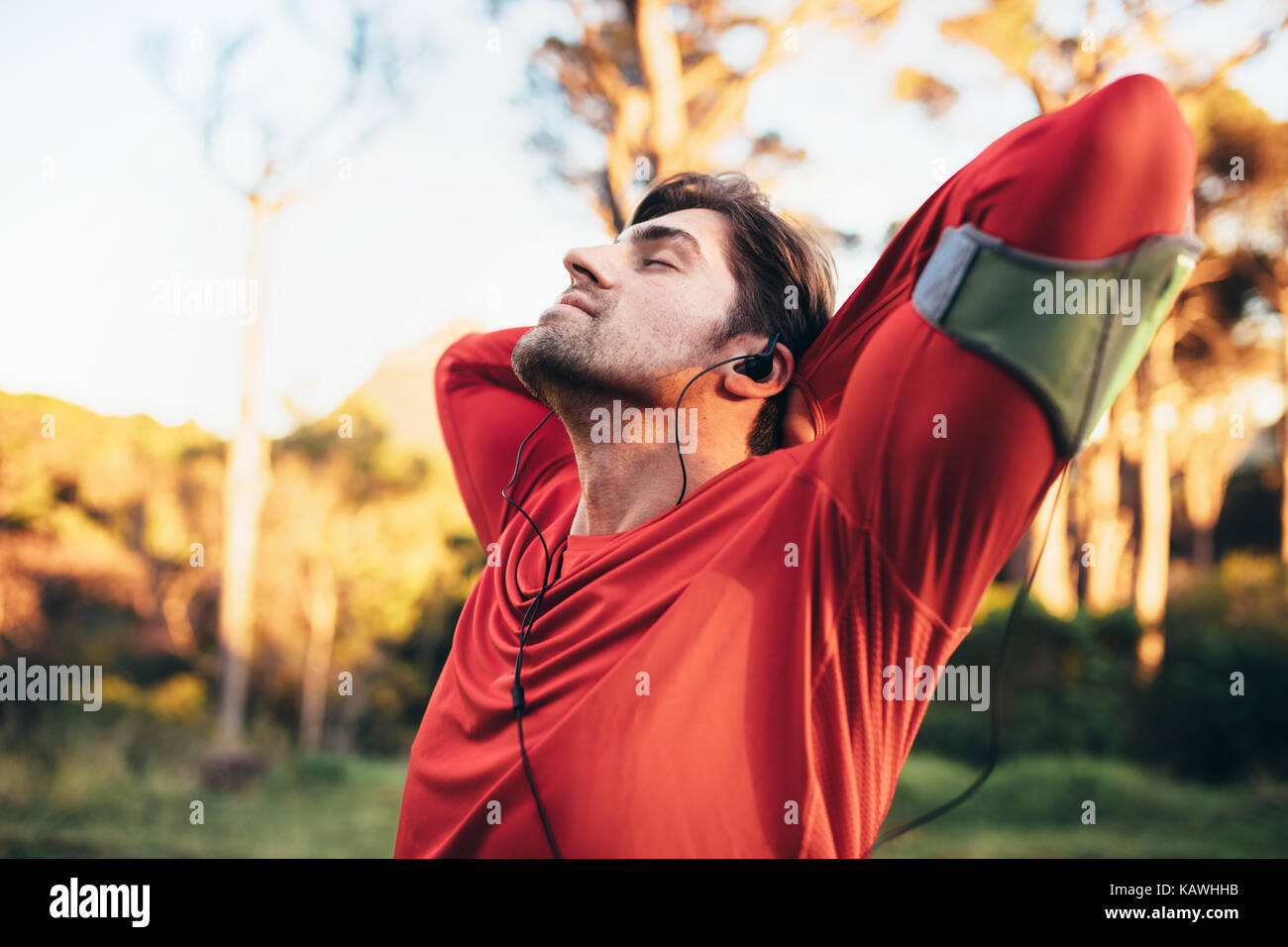 Runner doing breathing exercises while listening to music in a park. Man relaxing with closed eyes after workout. Stock Photo