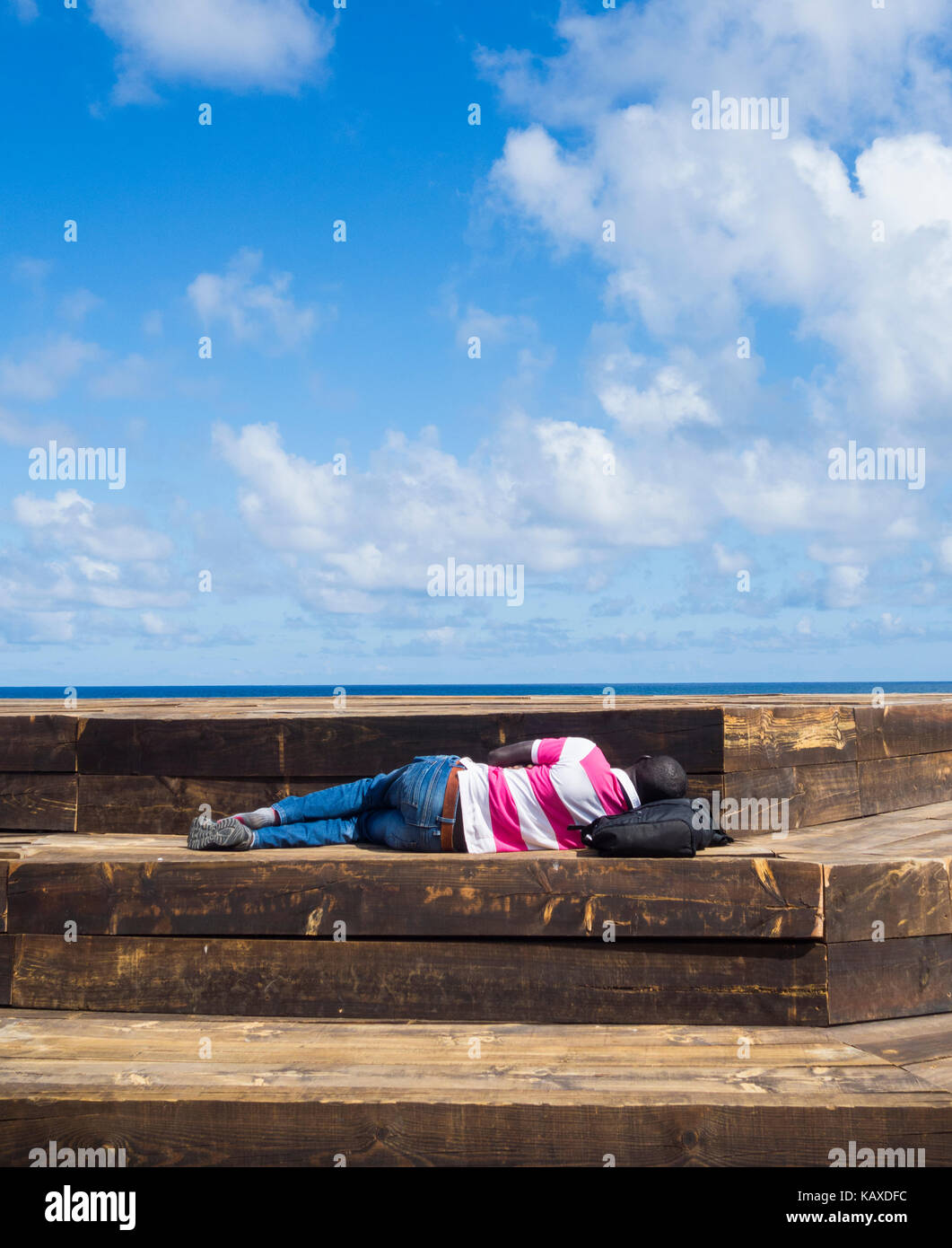 African man sleeping on wooden seat overlooking the sea in Spain Stock Photo