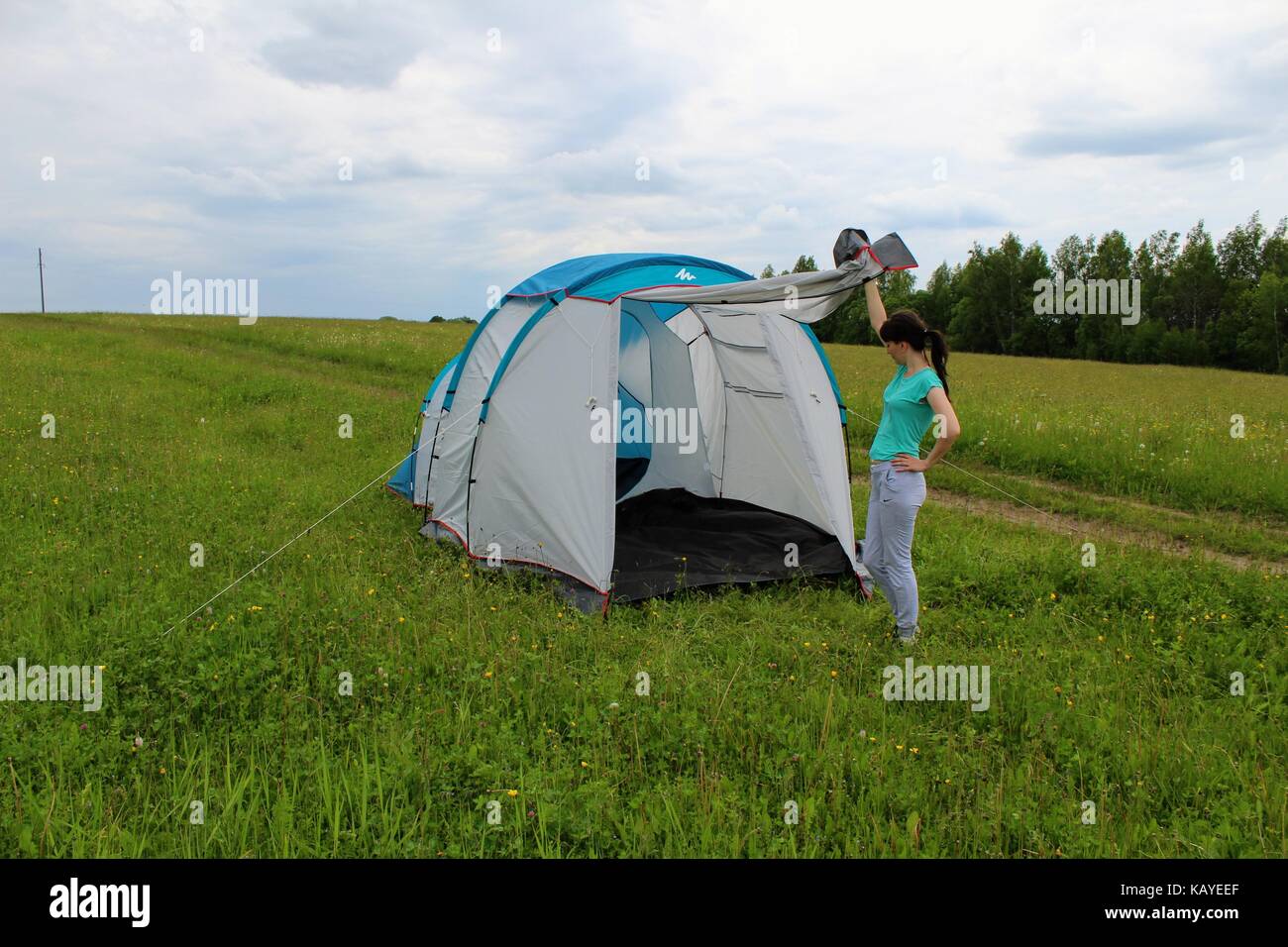 A young girl lifts the canopy of a white-blue tourist tent set on a green meadow against a forest background. Stock Photo