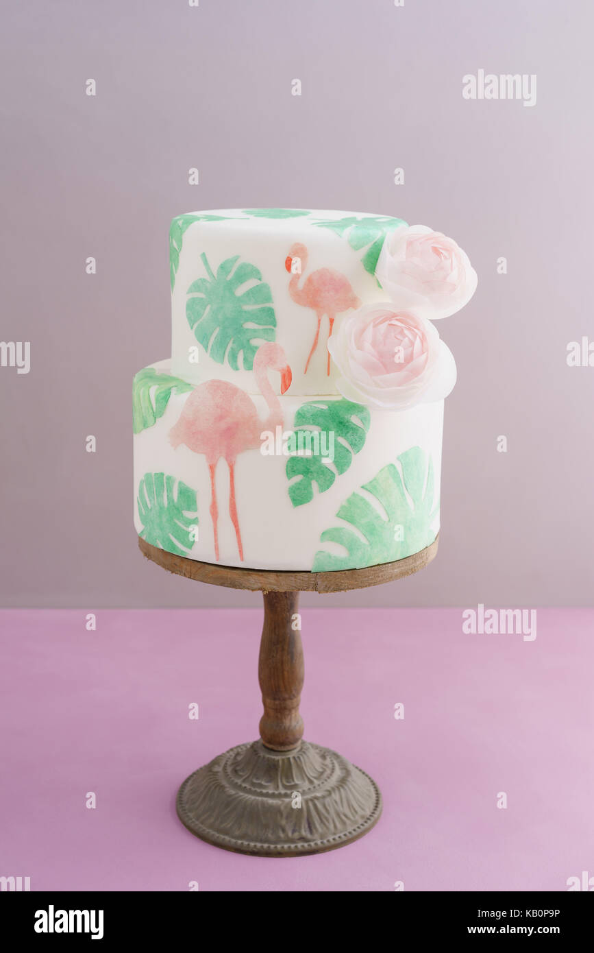 Two tiered tropical wedding cake with fondant, tropical wafer paper leaves and flamingos with ranunculus flowers on wooden cake stand Stock Photo