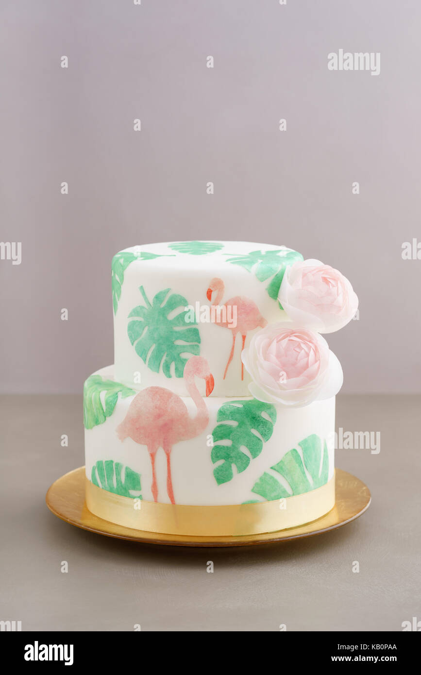 Two tiered tropical wedding cake with fondant, tropical wafer paper leaves and flamingos with ranunculus flowers on golden cale platter Stock Photo