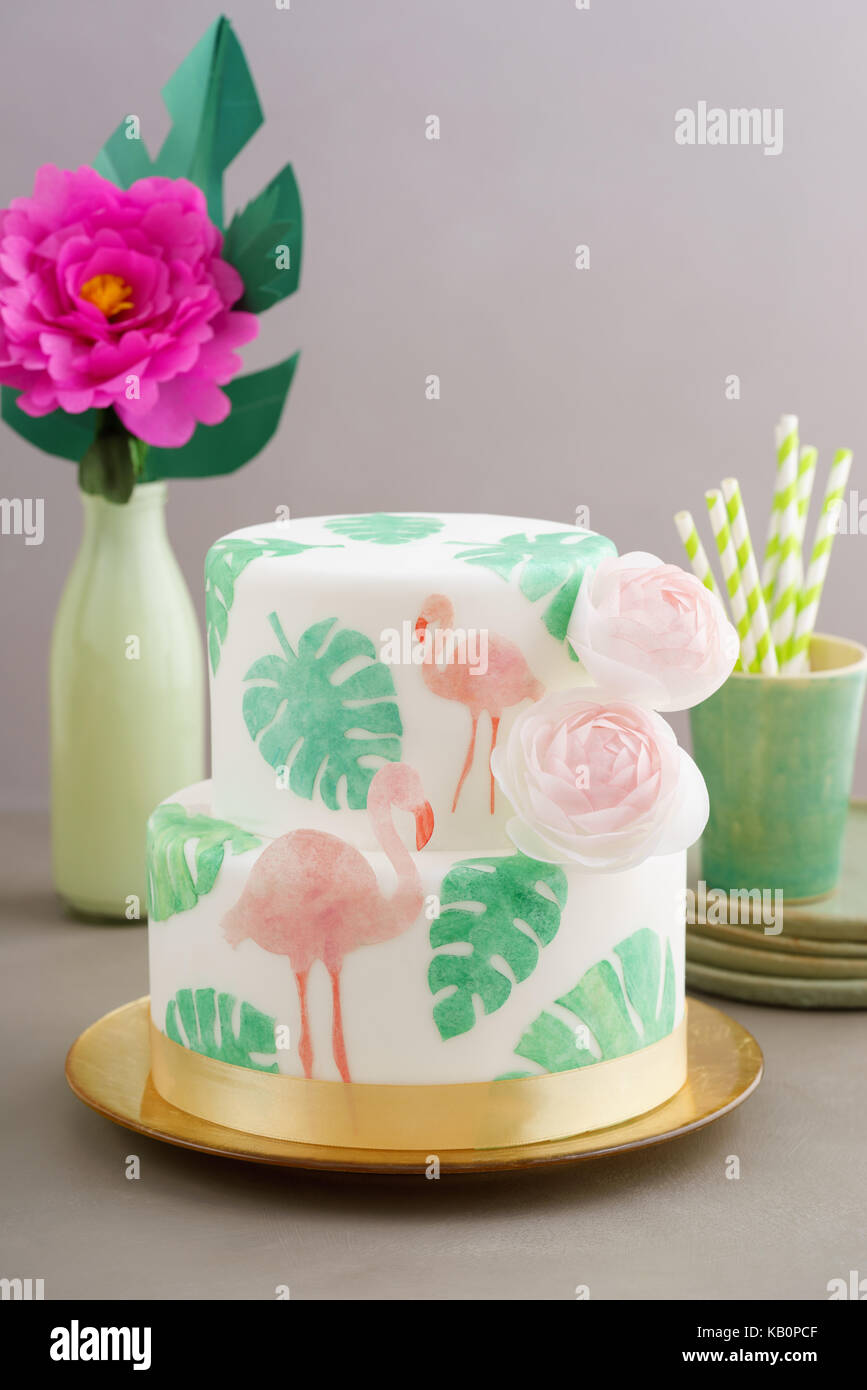 Two tiered tropical wedding cake with fondant, tropical wafer paper leaves and flamingos with ranunculus flowers on golden cale platter Stock Photo