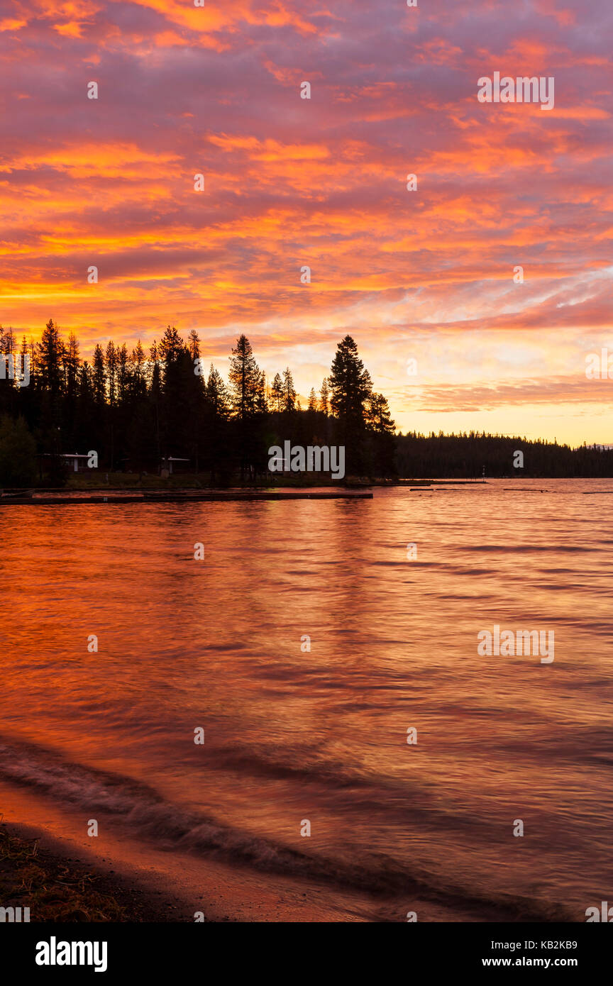 Peaceful and colourful red sky sunrise at Diamond Lake with waves splashing on the shore and pine tree silhouettes on the far shore, Oregon, USA. Stock Photo