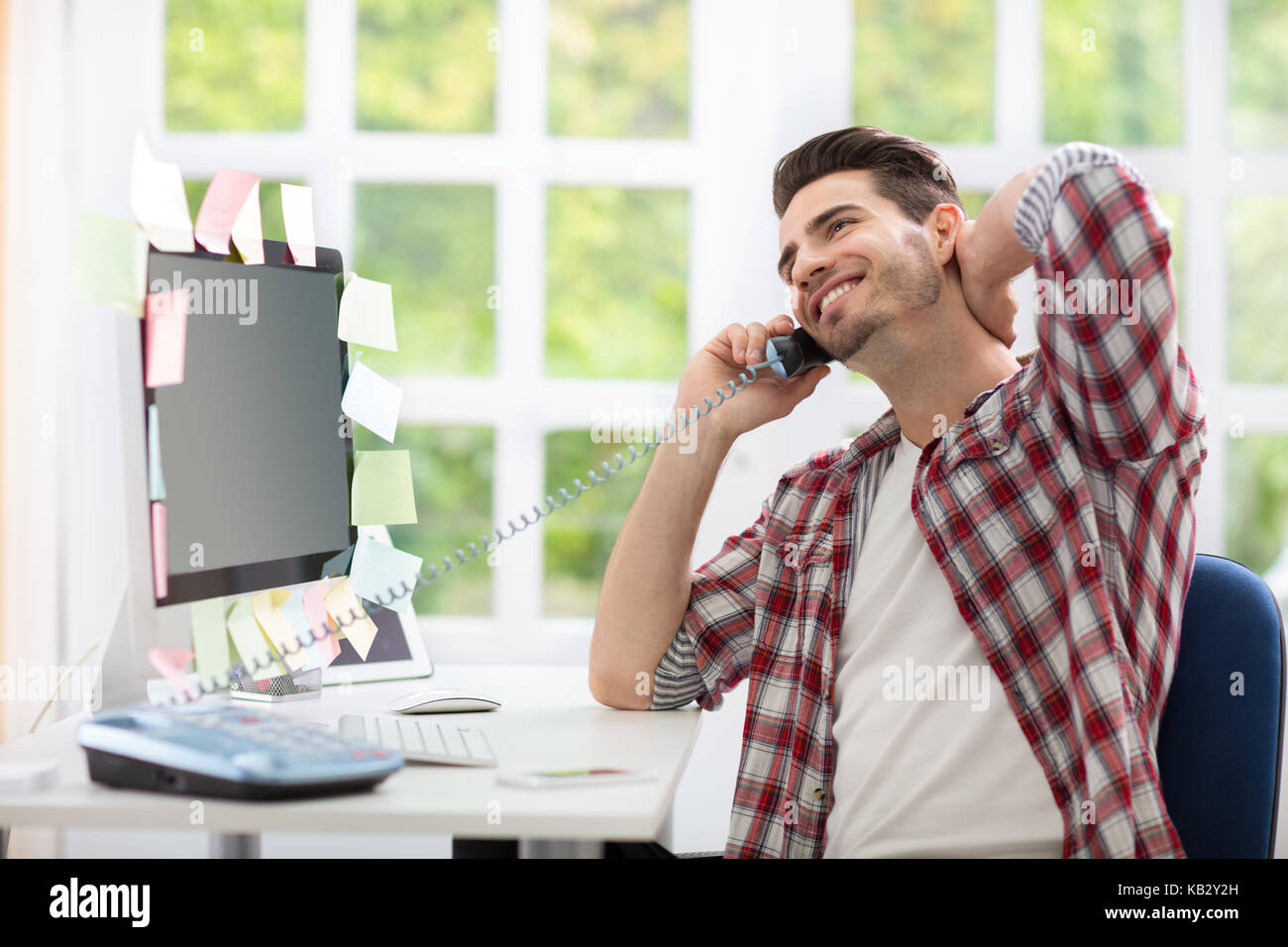 Cheerful young man sitting at work desk front of computer and  talking on phone Stock Photo