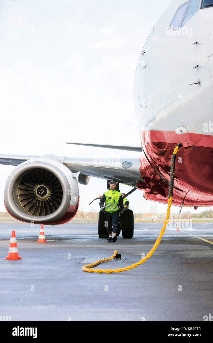 Full length of ground crew member walking while airplane being charged on runway against sky Stock Photo