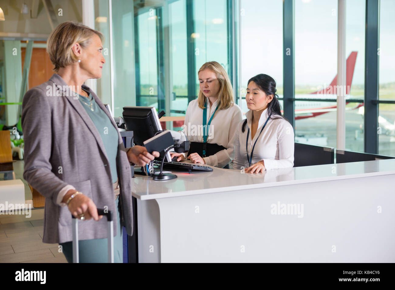 Female passenger holding passport while looking at receptionists working at counter in airport Stock Photo