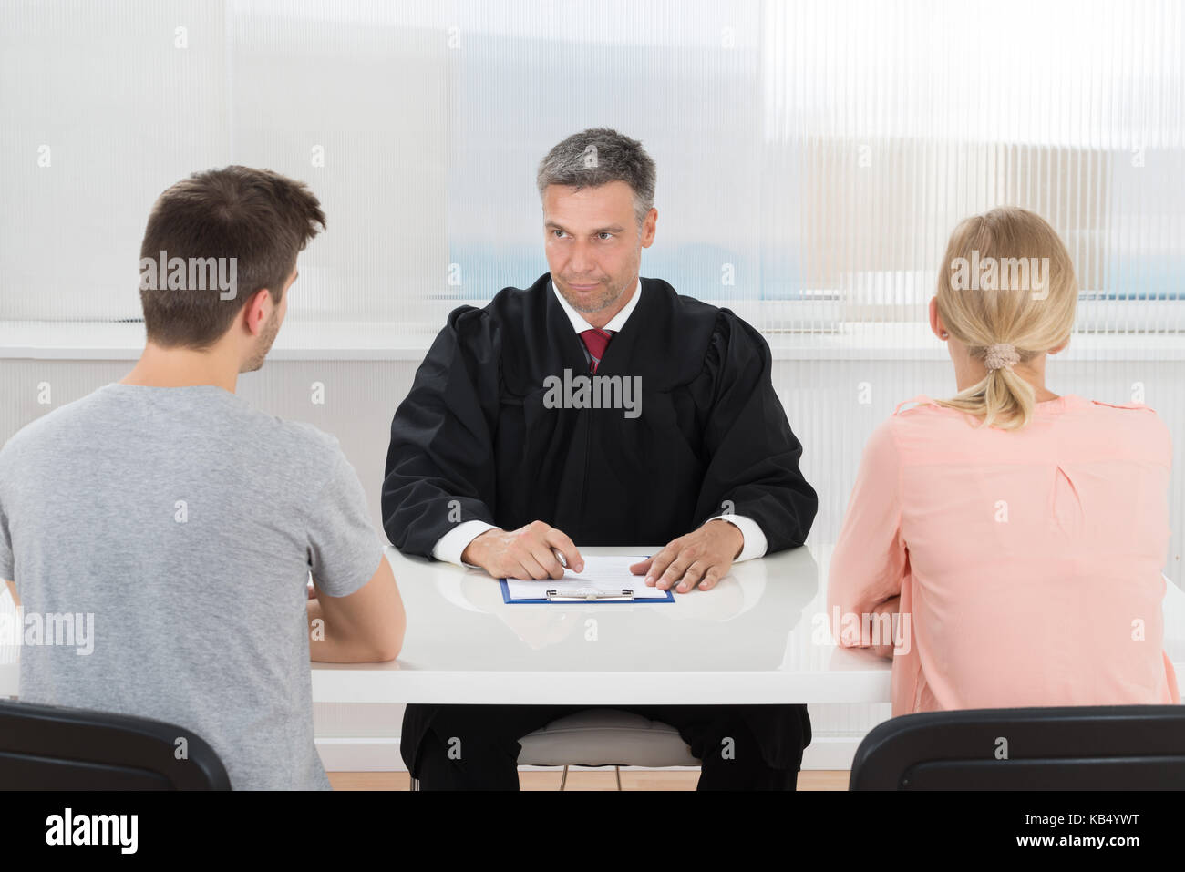 Mature Male Judge Sitting In Front Of Young Couple In Courtroom Stock Photo