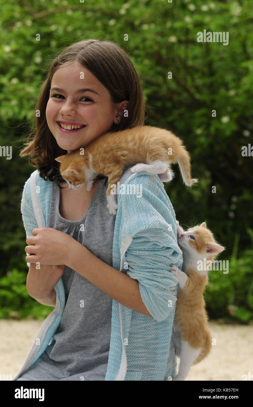 Girl with two kittens on shoulder and back laughs in the camera, trees in the background, blurred Stock Photo