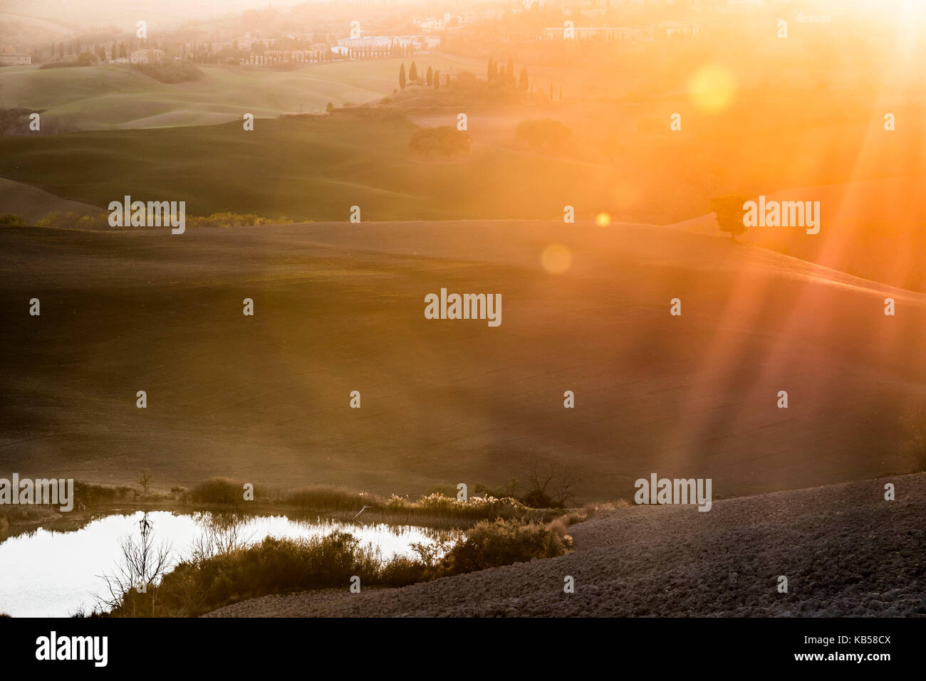 The last rays of setting sun and sun flares are touching a small pond and gray Tuscany ground Stock Photo
