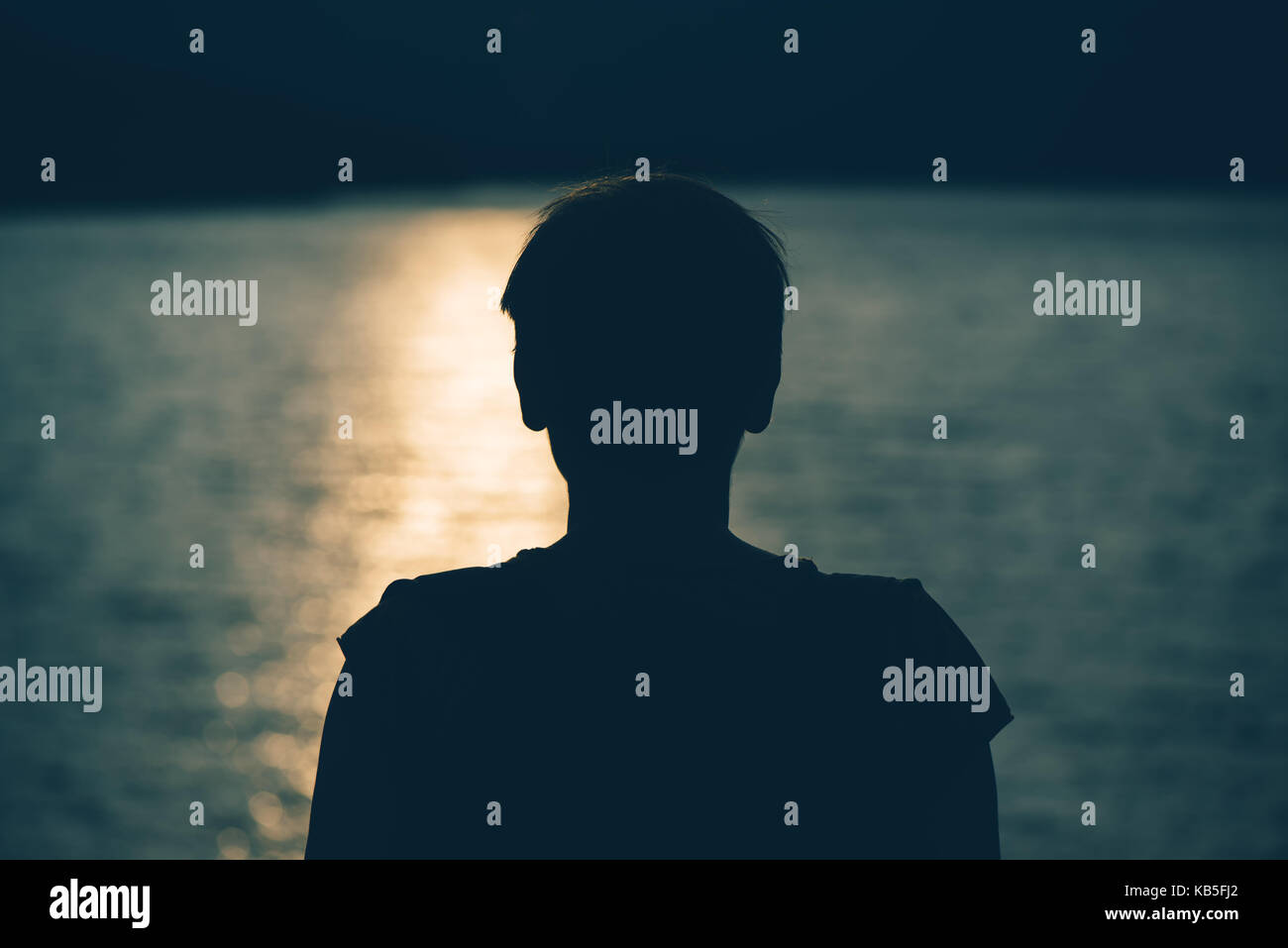 Silhouette of depressed sad woman standing by the lake in cold autumn sunset. Gloomy adult female person looking at water. Loneliness and solitude con Stock Photo