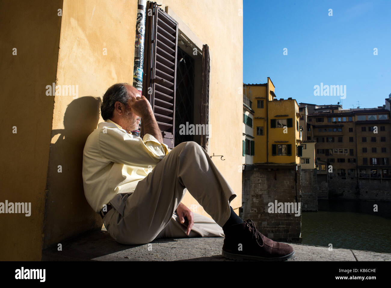A man sits thoughtfully on the Arno river bank with Ponte Vecchio bridge on the back. Florence, Italy Stock Photo