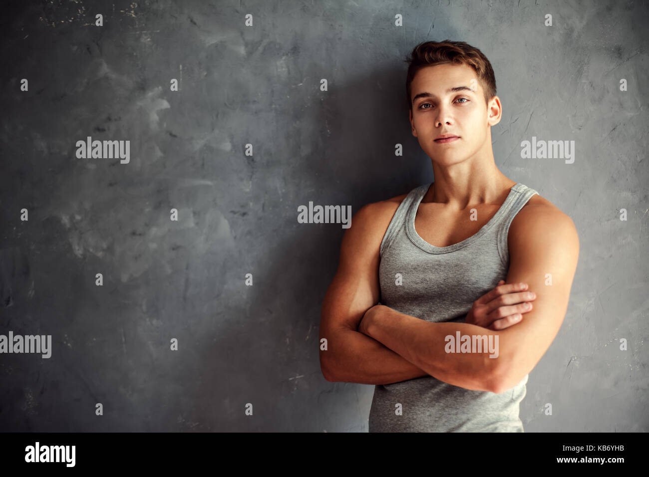 Portrait of muscular young handsome man wearing a gray undershirt standing with hands folded against gray textured wall Stock Photo