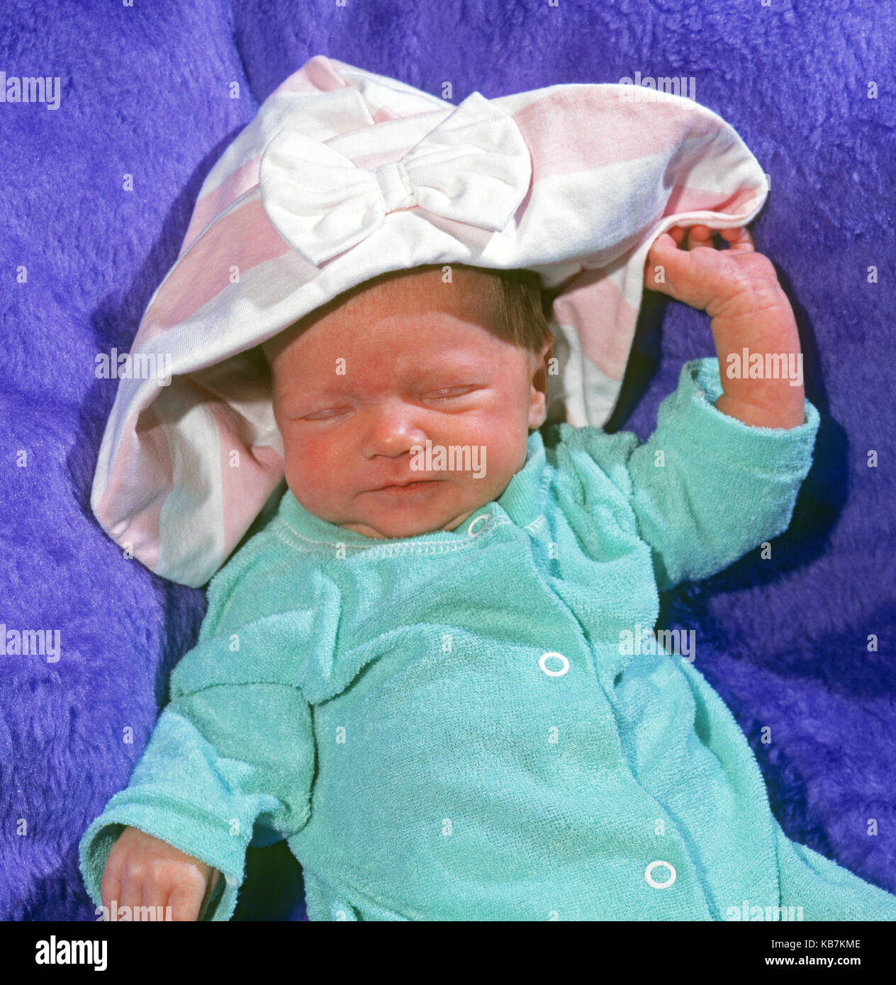 A two-week old baby wearing a onesie and a bonnet taking a nap in a blue blanket. Stock Photo