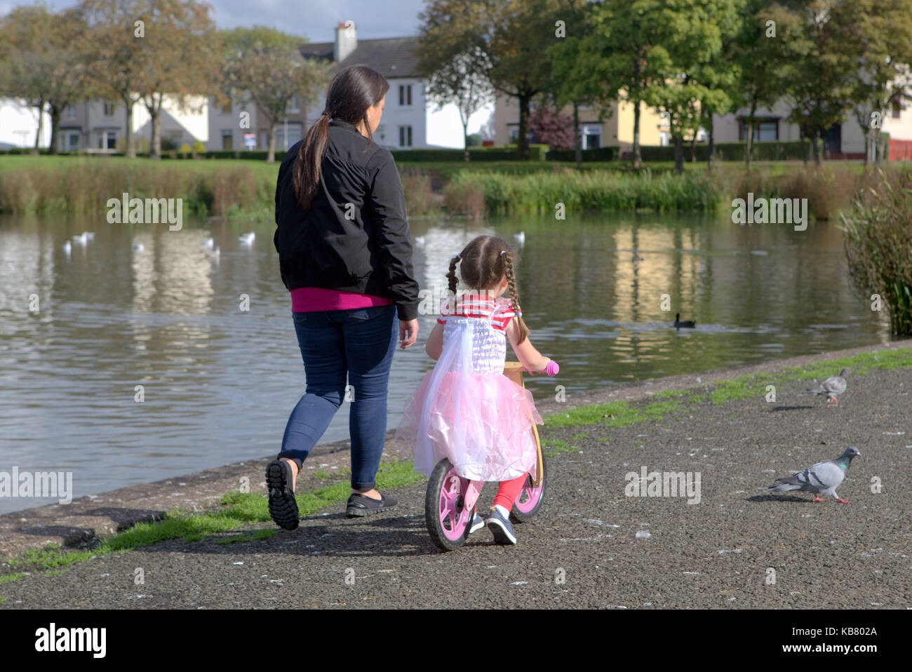 little girl dressed as a princess on a balance bicycle with her mother sunny day beside pond Glasgow Stock Photo