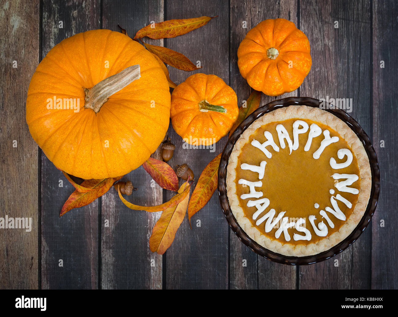 Pumpkin pie with Happy Thanksgiving text. Displayed with pumpkins, golden autumn leaves, and acorns on rustic table. Top view. Stock Photo